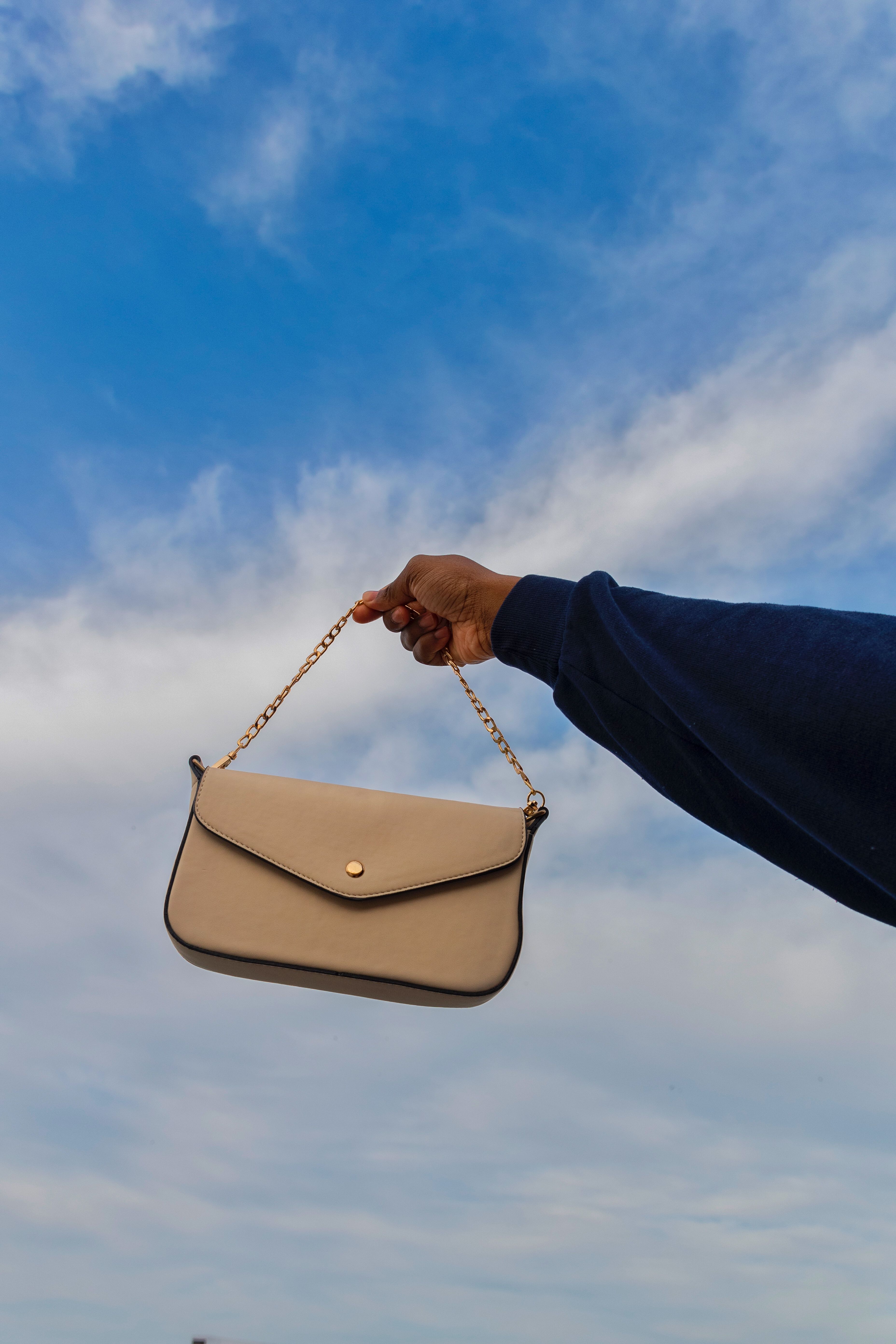 Beige crossbody handbag with a gold chain strap against a blue sky